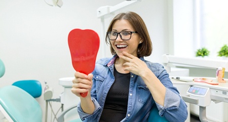 A woman smiling at her new denture in the mirror at the dentist office